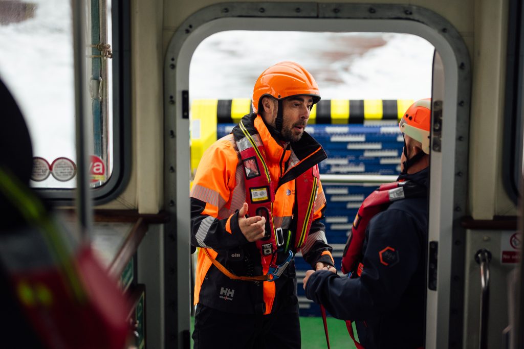 Two crew members in safety gear discuss operations next to the SWIFT recovery system aboard the pilot boat.