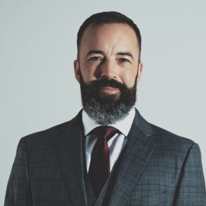 Portrait of Commander Matthew Mitchell, wearing a dark checkered suit, maroon tie, and white shirt, with a neatly groomed beard and confident expression.