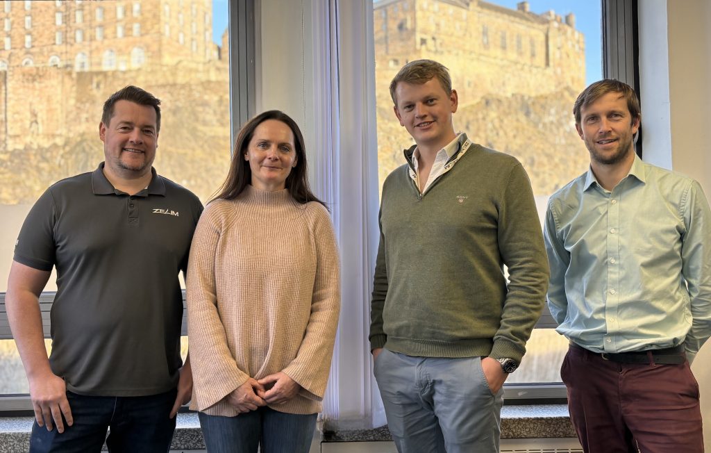 A group photo of Zelim's leadership team and investor Ali Clark from Amati. From left to right: Alan Gilbert, CFO, wearing a grey polo; Ali Clark, wearing a pink sweater; Sam Mayall, CEO, in a green jumper; and Andy Tipping, Business Development Director, in a light blue shirt. Edinburgh Castle is visible through the window in the background.