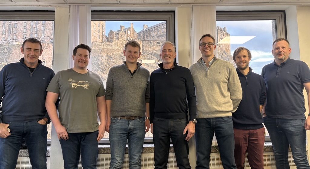 A group of seven men from Zelim's leadership team stand together in an office with Edinburgh Castle visible through the windows behind them. They are dressed in smart casual attire and smiling at the camera.